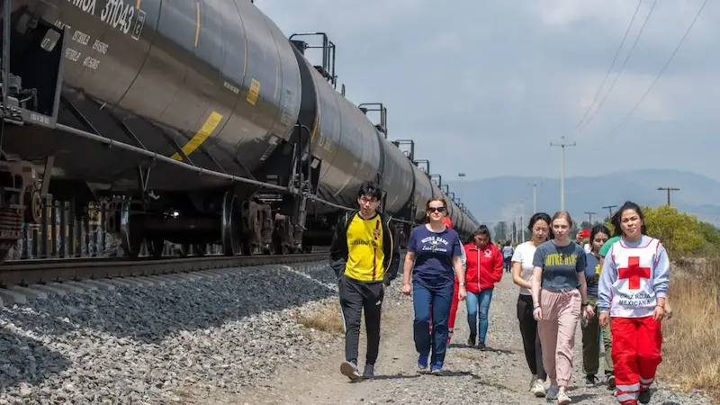 A group of people walks alongside railroad tracks with large train cars on the left and a dry, grassy field on the right.